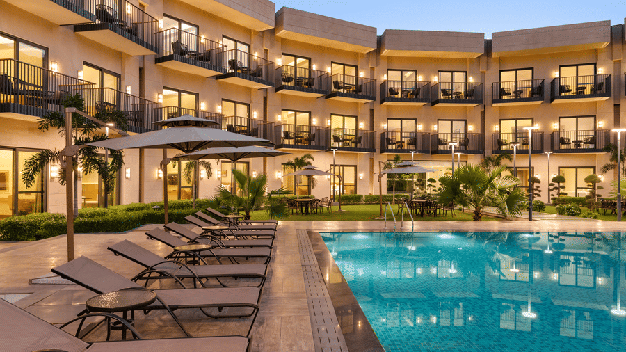 Stunning outdoor swimming pool at Ramada by Wyndham Sonipat Murthal, surrounded by sun loungers, lush palm trees, and the illuminated multi-storey hotel facade at dusk.