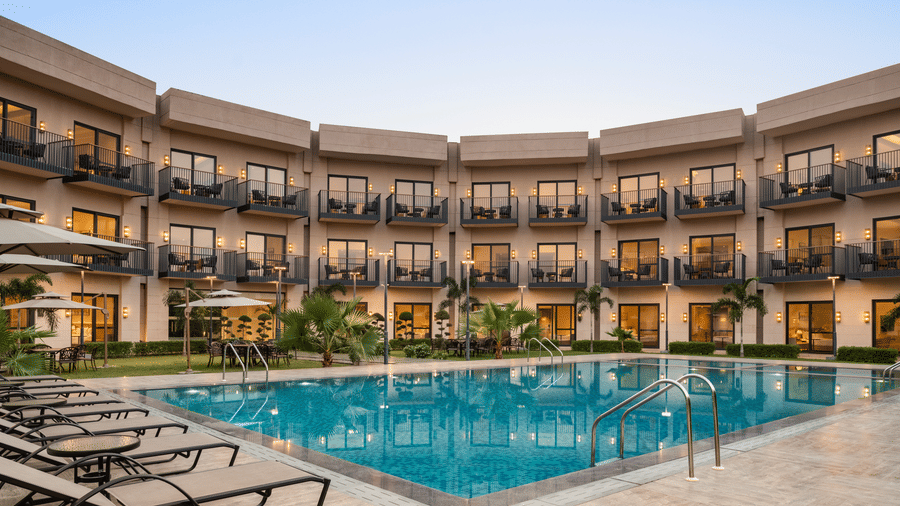 Panoramic view of the outdoor pool area at Ramada by Wyndham Sonipat Murthal, with sun loungers along the poolside, tropical landscaping, and the hotel's curved balconied facade at sunset.