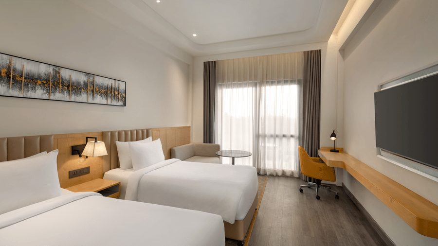 Twin-bed hotel room with 2 single beds, wooden headboards, a large flat-screen TV, and floor-to-ceiling curtained windows.