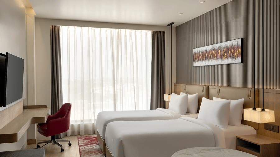 Spacious hotel room with twin beds dressed in white linens, a red accent chair, sheer curtains, and a wall-mounted flat-screen television.