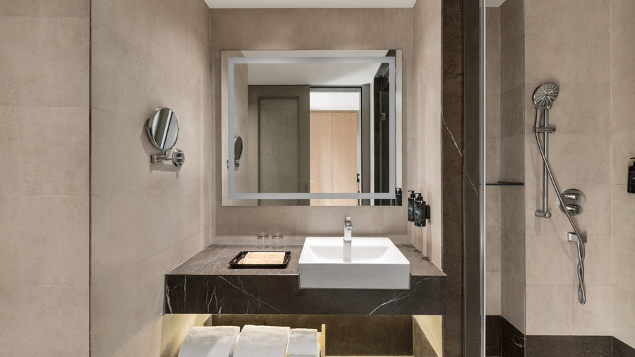 Elegant hotel bathroom corridor with a long vanity mirror, vessel sink, wooden cabinetry, and a glass-enclosed shower area in neutral tones.