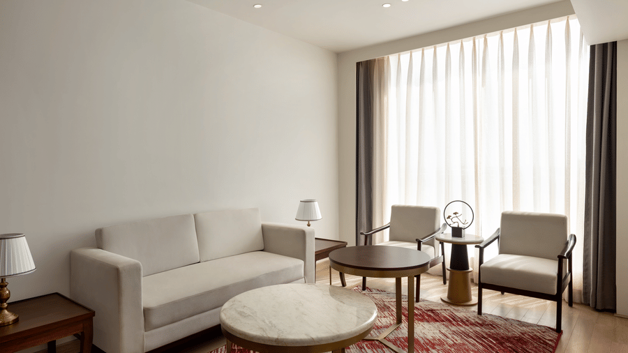 Bright and airy suite living area with a white sofa set, armchairs, a round coffee table, and large floor-to-ceiling curtained windows.