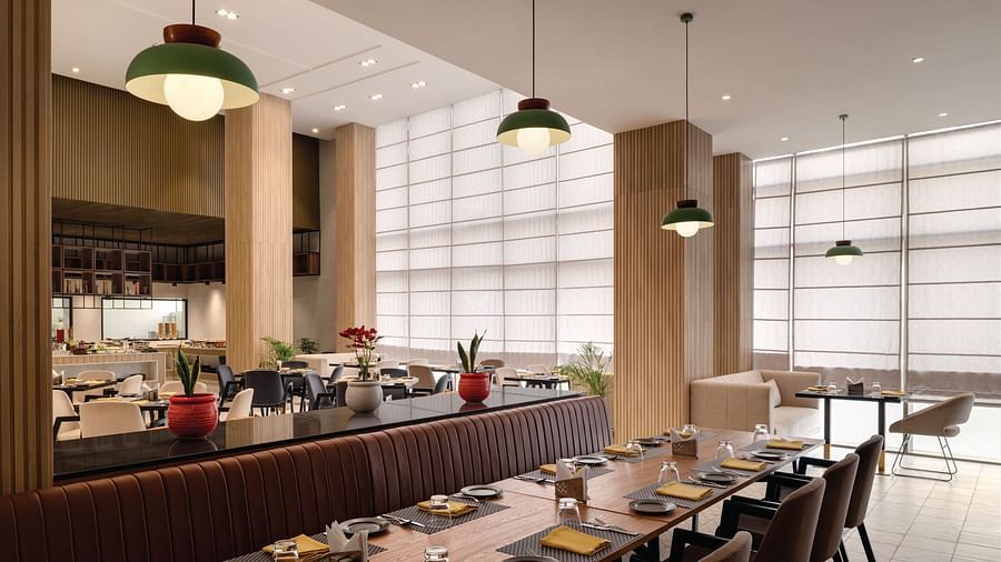 Ramada Encore by Wyndham, Bhiwadi | Restaurant with a long table, pendant lights, and a view of the kitchen.