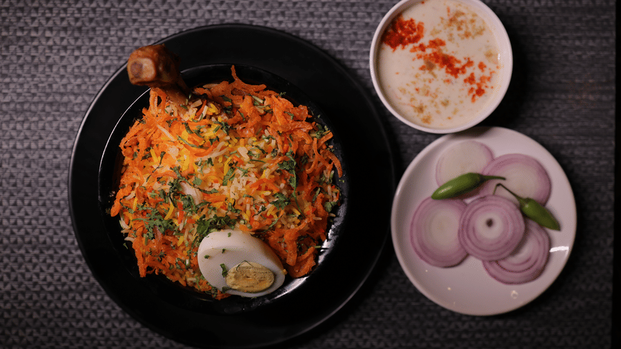 A plate of traditional chicken biryani served with a side of creamy raita, sliced onions and green chillies.
