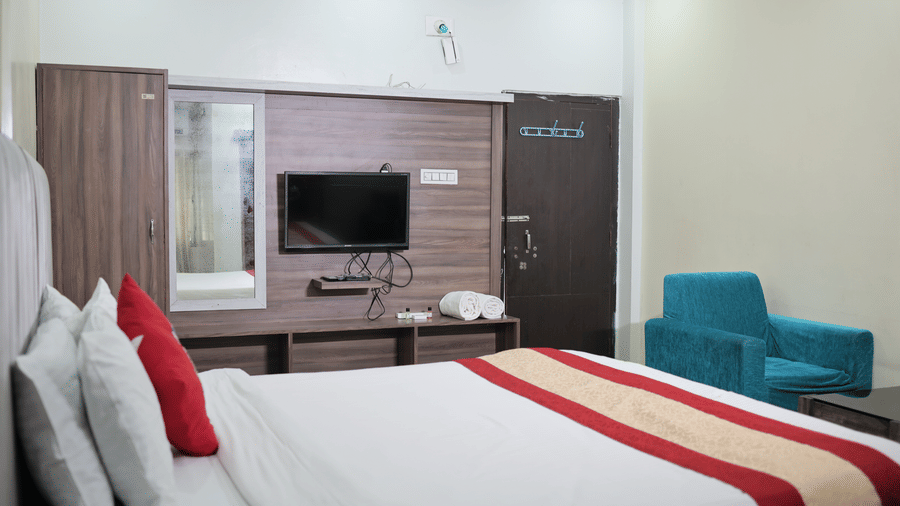 A well-lit hotel bedroom showing a neatly made double bed, a wall-mounted television, and a blue armchair.