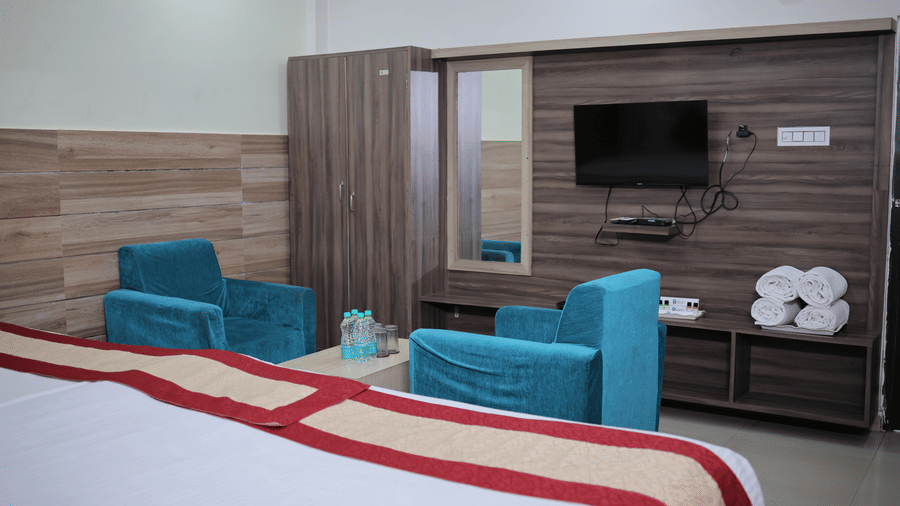 A hotel room interior featuring two bright blue armchairs, a wooden wardrobe, and a wall-mounted television.