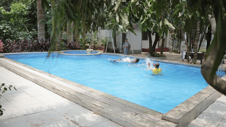 People enjoying a swim in a clear, blue outdoor swimming pool surrounded by green trees.