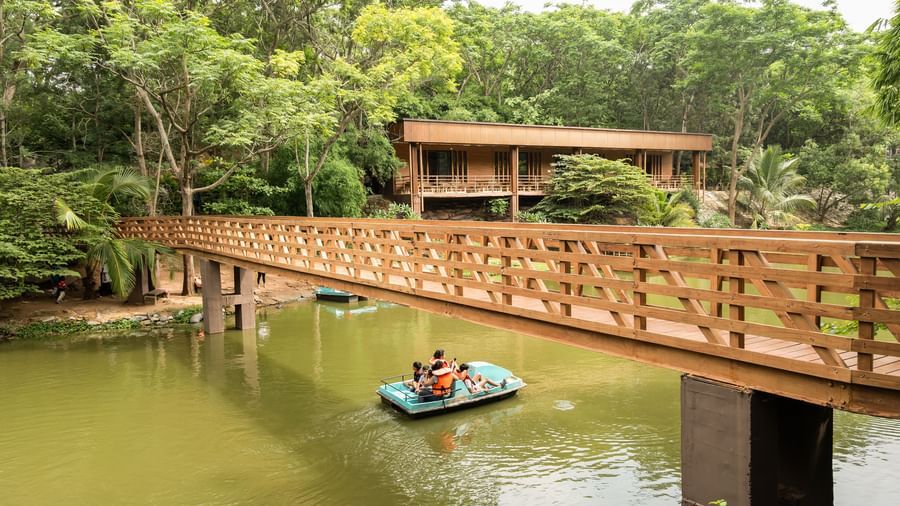 A wooden bridge spans across a calm body of water where a small boat is visible, surrounded by abundant green foliage | Lake View Restaurant