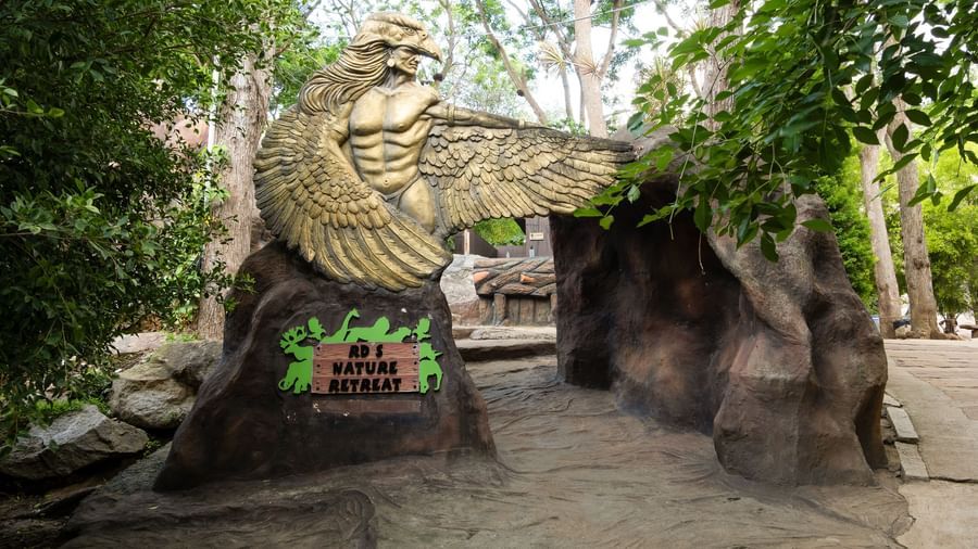 A large stone eagle sculpture stands at the entrance of a lush, green pathway leading into a natural-themed area