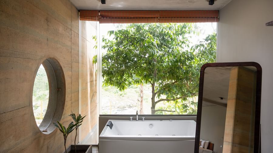A bathroom featuring a large round mirror, a vanity area, and a window overlooking lush trees | Gaia Studio | RD's Nature Retreat, Bangalore
