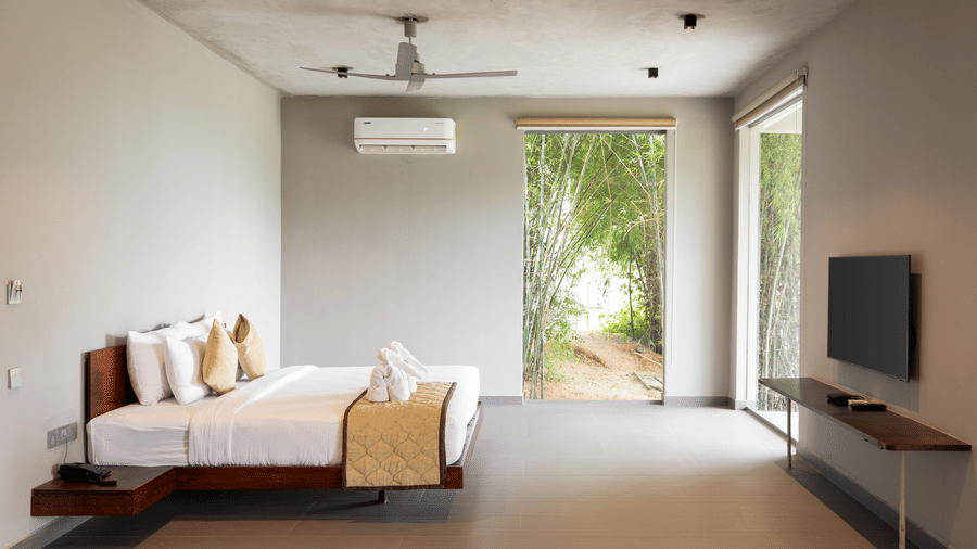 A minimalist bedroom of Gaia Studio with a large bed in front of a television, and an open doorway leading to an outdoor area at RD's Nature Retreat, Bangalore