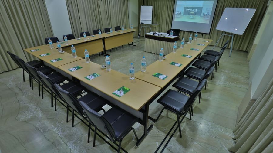 An empty conference room set up with tables in a U-shape, chairs, a projector screen, and a whiteboard - Retreat Heritage, Lonavala
