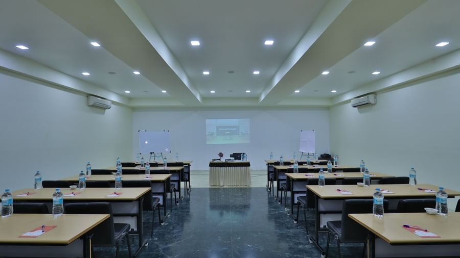 A spacious room with rows of tables and chairs facing a front presentation area with a screen - Retreat Heritage, Lonavala
