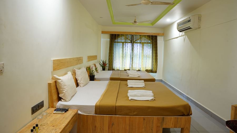 A side view of a room at Riverside Inn, featuring two beds placed beside each other, a bedside table, windows with curtains at the end, and a wall-mounted AC.