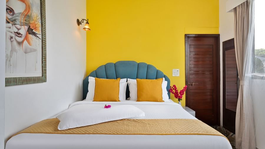 A neatly arranged bed with mustard and white linen, a teal headboard, and a yellow accent wall complemented by artwork.