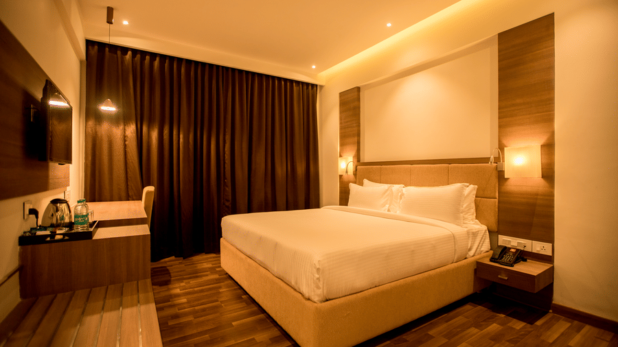 A wide view of a modern hotel bedroom showing a king size bed, bedside tables, and dark curtains illuminated by warm light at S Hotels, Chennai.