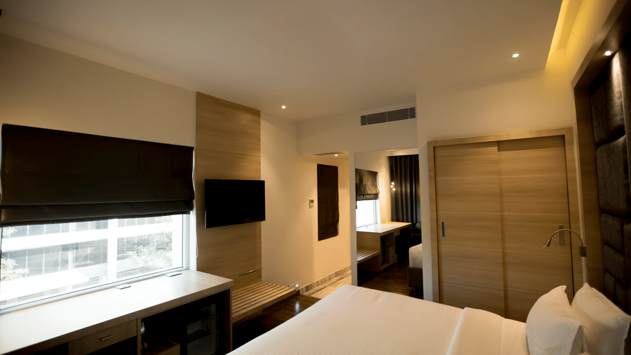 A hotel room features a king size bed with a dark headboard and a large window providing daylight into the room at S Hotels, Chennai.