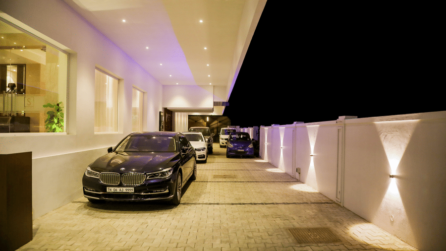 A dark coloured car is parked at the end of a brightly lit covered driveway leading into a building at night at S Hotels, Chennai.