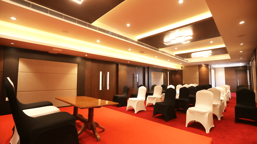 A banquet hall with a bright red carpet is set up with a table and chairs covered in black cloth at S Hotels, Chennai.