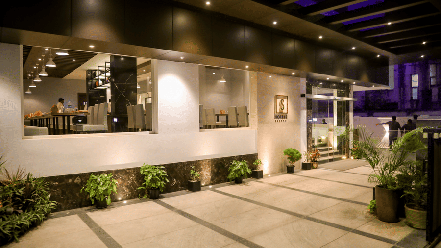 A modern building entrance features large glass windows, light coloured paving and manicured plants outside the front at S Hotels, Chennai.