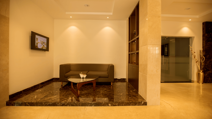 A small hotel lobby area contains a low wooden sofa, a coffee table and light beige walls with recessed lighting at S Hotels, Chennai.