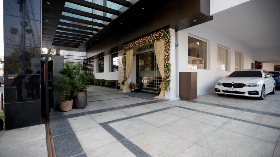 A car is parked in a modern outdoor covered portico area with large stone floor tiles and plants at S Hotels, Chennai.