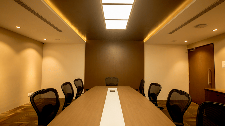 A long oval conference table with black chairs sits beneath a modern ceiling with strip lighting in the boardroom Pluto at S Hotels, Chennai.