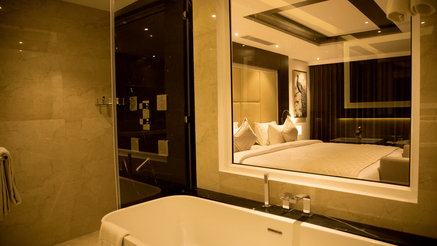 A large white bathtub is visible in a hotel bathroom with light brown tiling and a mirror reflecting the room at S Hotels, Chennai.