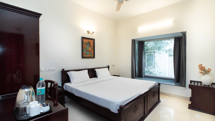 A room in Sanctum Suites Indiranagar, Bangalore, showcasing a double bed with white bedding, a bedside table with a lamp, an electric kettle placed on a table, a chair, and a window providing natural light.
