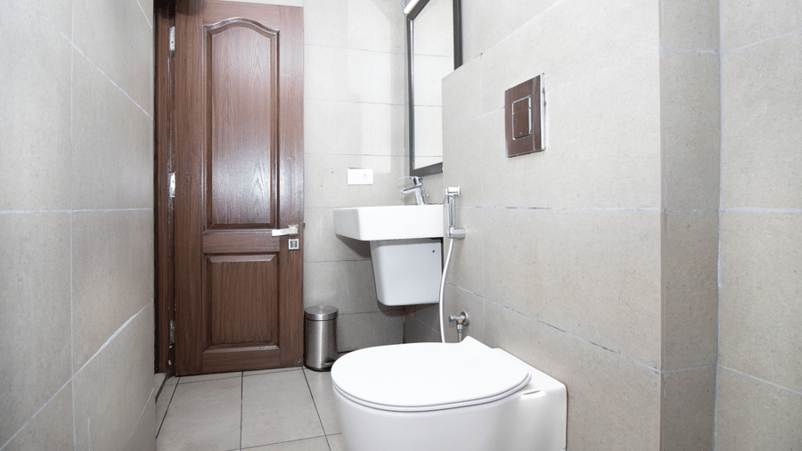 A bathroom at Sanctum Suites, Richmond Road, Bangalore includes a white toilet, a washbasin, a mirror, and a dark wood door.