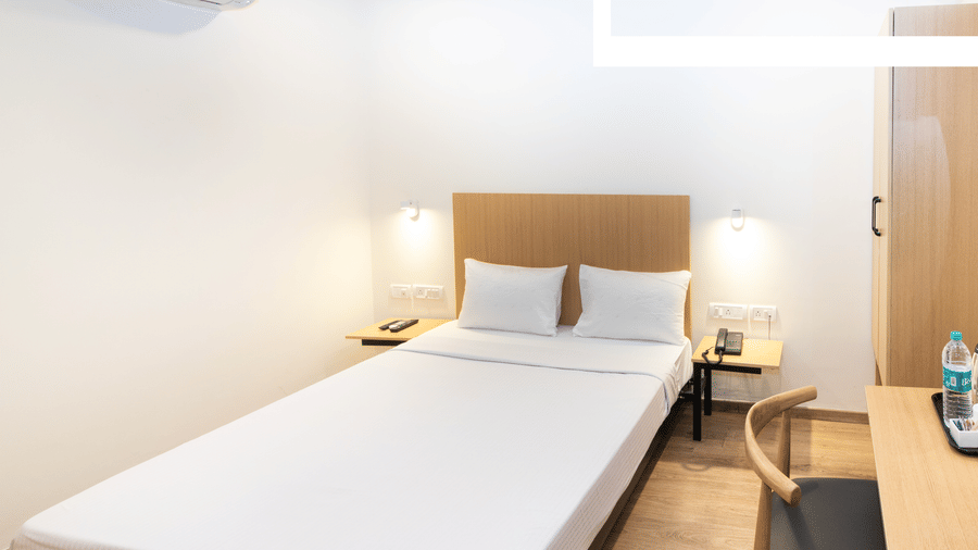A room at Sanctum Suites, Richmond Road, Bangalore features a bed with white sheets, a light wood headboard, a small side table, and a desk with a black chair.