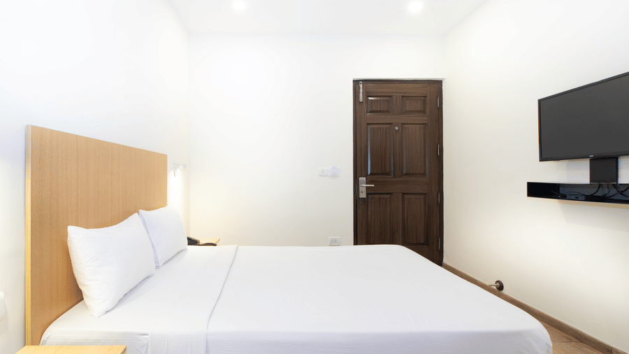 A room at Sanctum Suites, Richmond Road, Bangalore features a large bed, a wooden headboard, a television, and a dark wood entrance door.