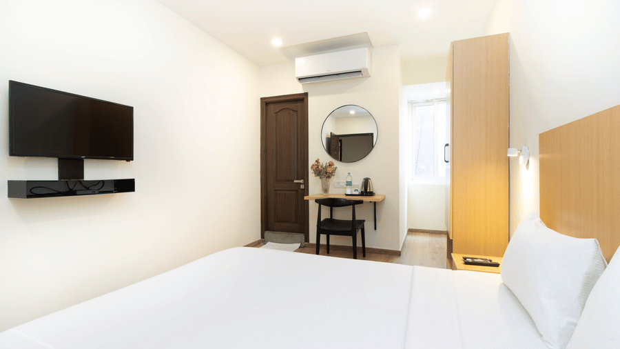 A bedroom at Sanctum Suites, Richmond Road, Bangalore features a double bed, a small dressing table with a round mirror, a wardrobe, a wall mounted A/C and a television.