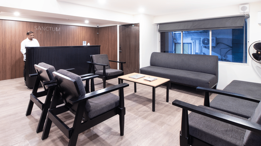 The lobby at Sanctum Suites, Richmond Road, Bangalore contains a dark reception desk, grey armchairs, a wooden coffee table, and a black sofa near a window with blinds.