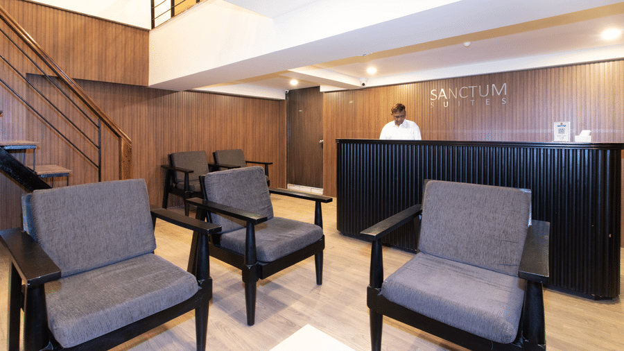 The lobby at Sanctum Suites, Richmond Road, Bangalore contains a dark reception desk, grey armchairs, and a wooden coffee table.