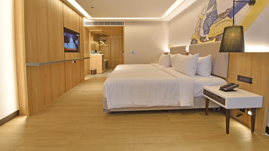 Modern bedroom at Sands Point Hotel with two beds, light wood paneling, a wall-mounted TV, and a colorful abstract mural behind the headboard. A bedside lamp and phone are visible.