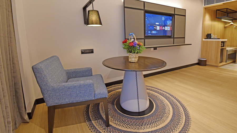 A seating area at Sands Point Hotel with a blue armchair, round wooden table, and flower vase set on a circular rug, featuring a wall-mounted TV in the background.