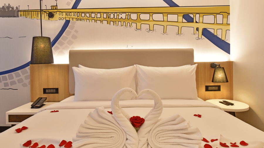 A bed at Sands Point Hotel decorated with swan-shaped towels and red rose petals. A bridge mural is on the wall behind the headboard between two bedside lamps.