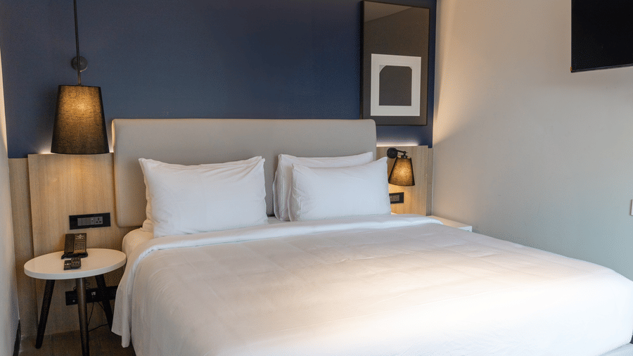 A Deluxe Suite king bedroom in the Sands Point Hotel, Pondicherry, featuring a large bed with a grey headboard, crisp white pillows and linen, a side-table under a black hanging lamp.