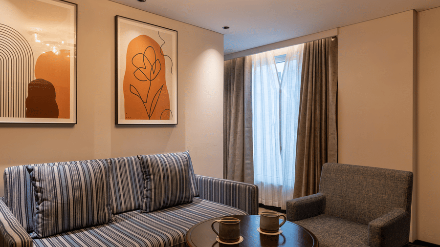 A Deluxe Suite living area in the Sands Point Hotel, Pondicherry, featuring a blue and white striped sofa with matching cushions, a grey armchair and a coffee table with 2 mugs kept on it.