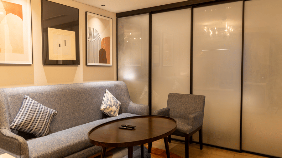 An Executive Suite in the Sands Point Hotel, Pondicherry, featuring a grey sofa with blue printed cushions, a round wooden coffee table, a grey armchair, 2 framed artworks mounted on the wall.