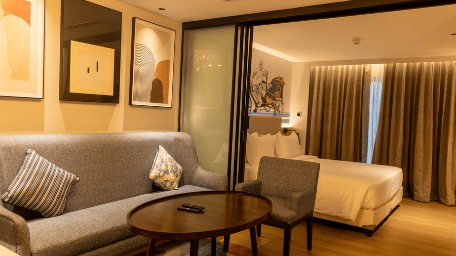 A wide-angle view of the Executive Suite at the Sands Point Hotel, Pondicherry, showing the layout with the living area featuring a grey sofa with cushions, a round dark coffee table, and framed artworks on the wall.