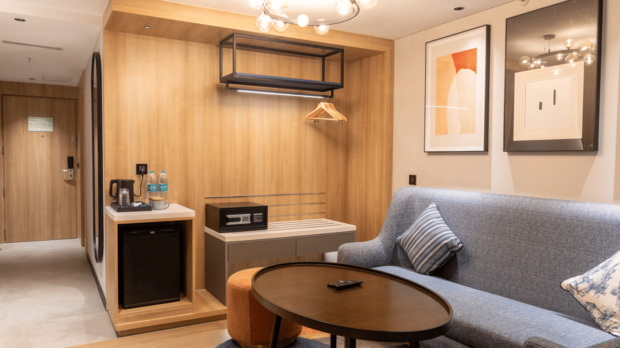 An in-room amenities corner in the Executive Suite at the Sands Point Hotel, Pondicherry, featuring a wardrobe with clothes hangers, a black digital safe, a minibar fridge,and a tea and coffee station with water bottles.