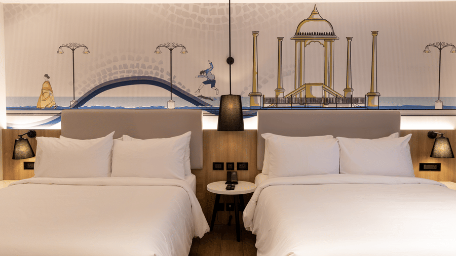 A close-up view of the 2 beds in the Family Room at Sands Point Hotel, Pondicherry, each in white linen with pillows and light grey headboards with warm integrated lighting, and shared bed lamps.