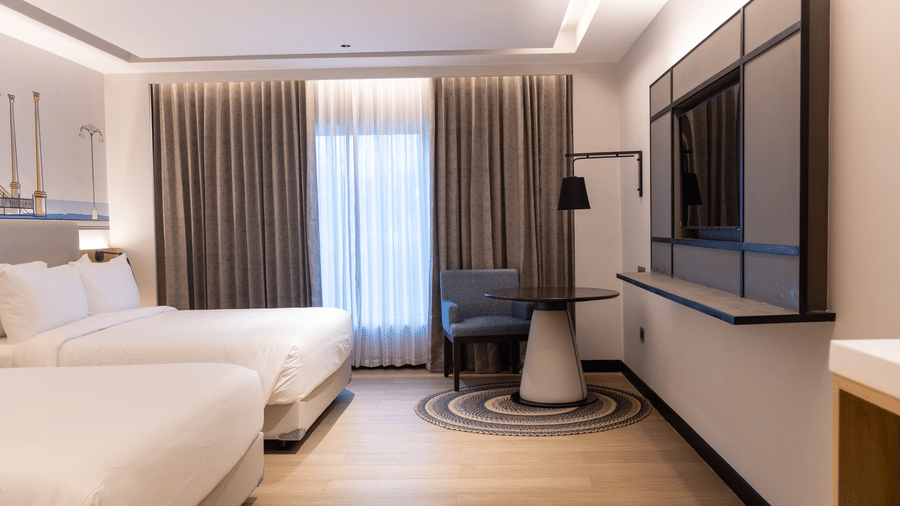 A Family Room at Sands Point Hotel, Pondicherry, featuring 2 beds with white linen, light grey headboards with built-in reading lamps, and a blue armchair on a circular braided rug. 