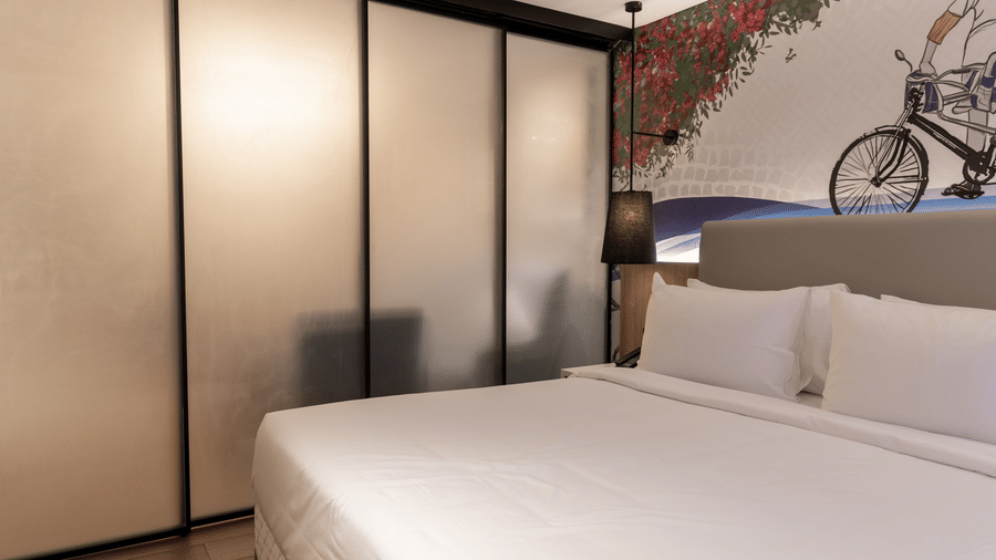 A bedroom in the Sands Point Hotel, Pondicherry, Luxury Suite featuring a king bed in white linen with a light grey headboard, a black pendant lamp, a full-height glass sliding partition with black frames separating the bedroom.