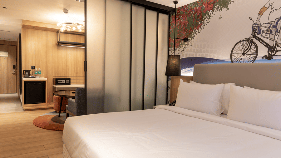 A wide view of the Sands Point Hotel, Pondicherry, Luxury Suite bedroom showing a king bed with white linen and a headboard, a black lamp, a frosted glass sliding partition.