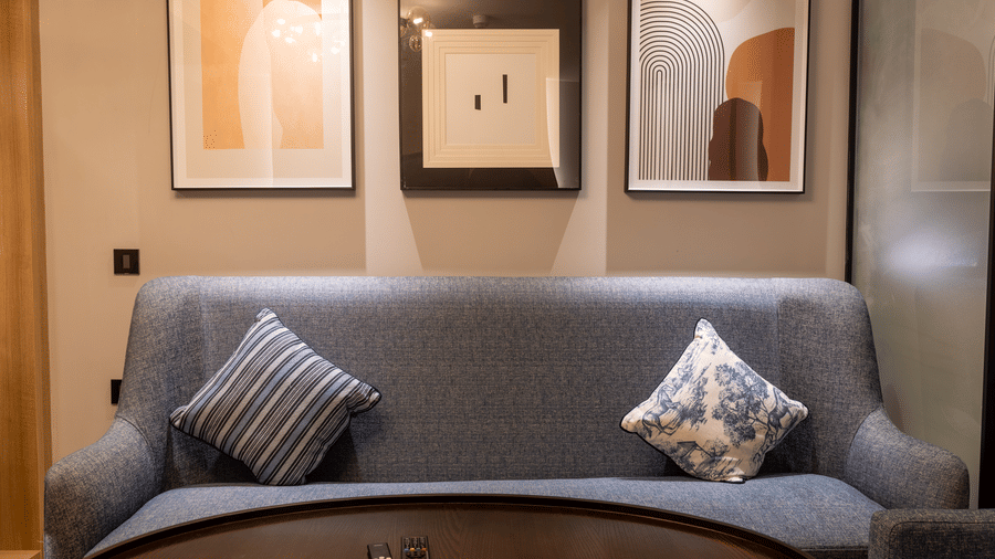 A living area in the Sands Point Hotel, Pondicherry, Luxury Suite featuring a curved grey sofa with patterned cushions, with a coffee table ans 3 artworks in the background.