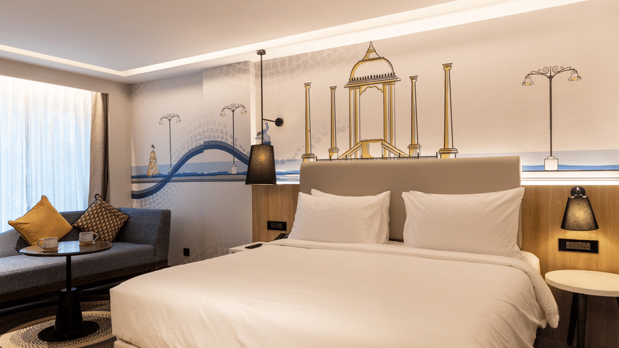 The Premium Room showcasing a king bed with white linen and a grey headboard with built-in reading lamps, and a grey sofa with yellow cushions, at the Sands Point Hotel, Pondicherry.