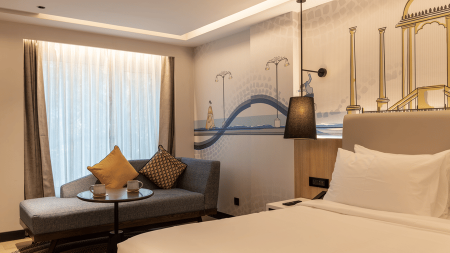The Premium Room showcasing a king bed with white linen and a grey sofa with yellow cushions, at the Sands Point Hotel, Pondicherry.